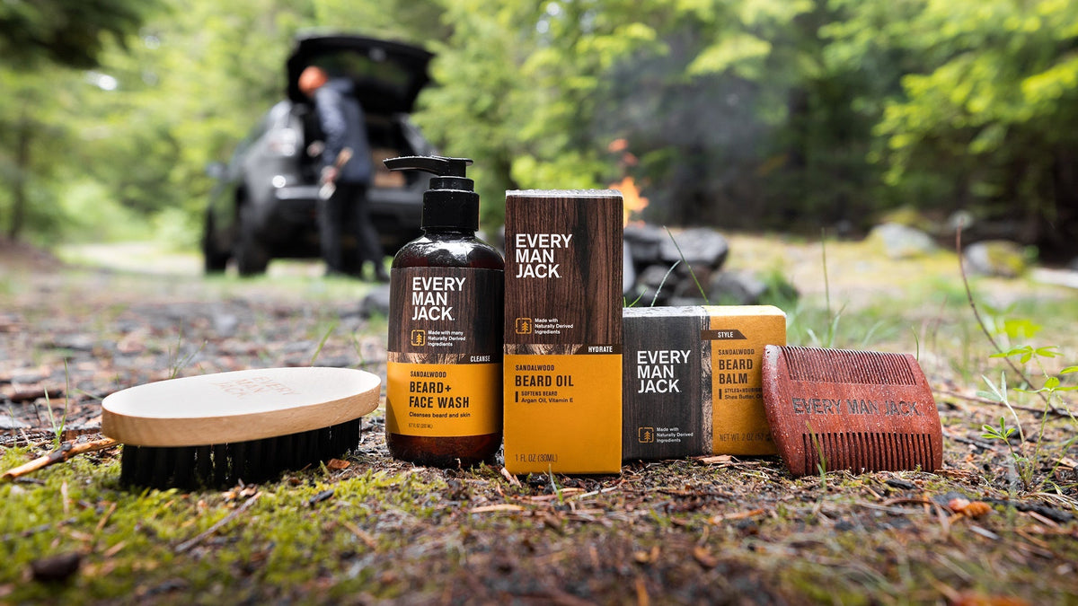 Every Man Jack beard-care products arranged on a mossy forest floor with wooden brush and comb, blurred car and campfire in the background