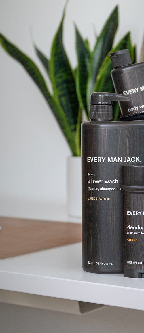 Grooming kit bundle with Every Man Jack body wash, deodorant, shampoo + conditioner, and face wash on a white desk with a backpack and plant in the background