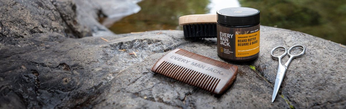 Beard grooming tools on a rock: wooden comb labeled 'EVERY MAN JACK', beard brush, jar of 'EVERY MAN JACK Beard Butter Sandalwood', and silver scissors.