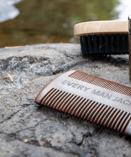 Beard grooming tools on a rock: wooden comb labeled 'EVERY MAN JACK', beard brush, jar of 'EVERY MAN JACK Beard Butter Sandalwood', and silver scissors.