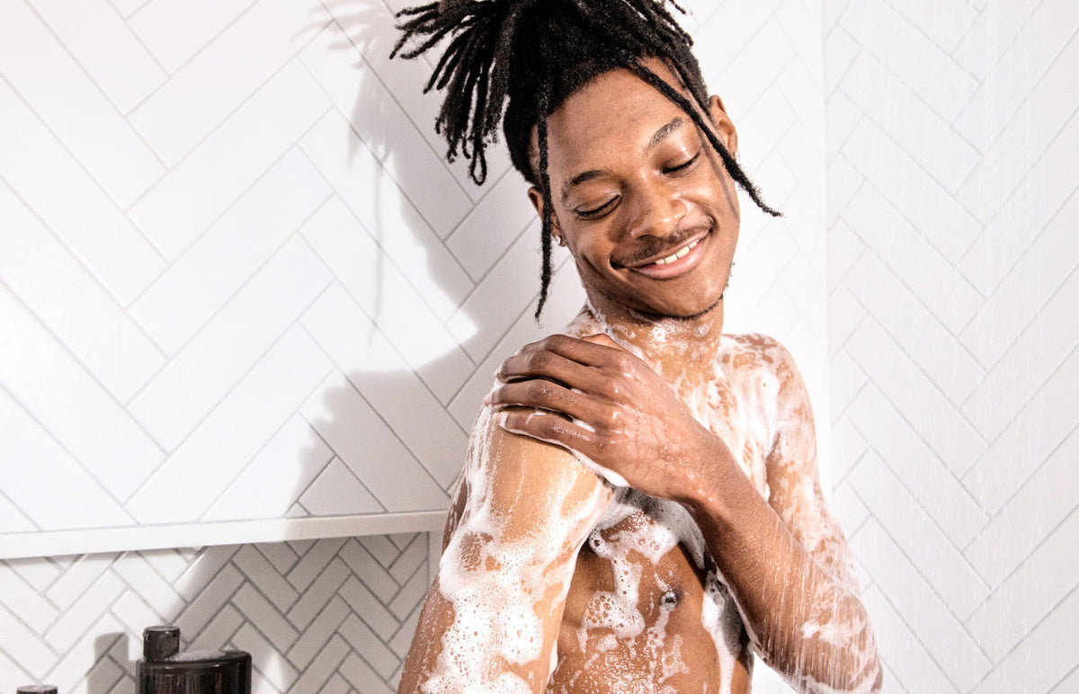 Man washing his shoulder with soapy lather in a white-tile shower, smiling