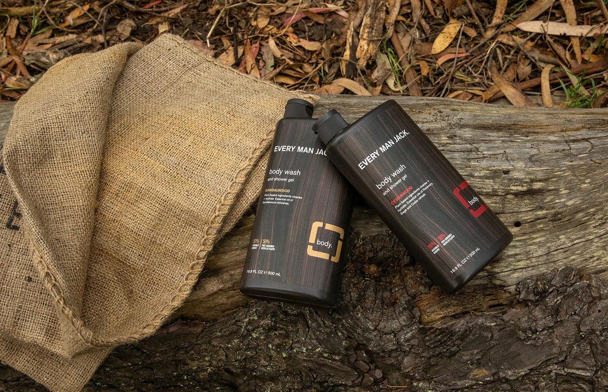 Two Every Man Jack body wash bottles (Sandalwood and Cedarwood) resting on a weathered log beside burlap in a rustic outdoor setting