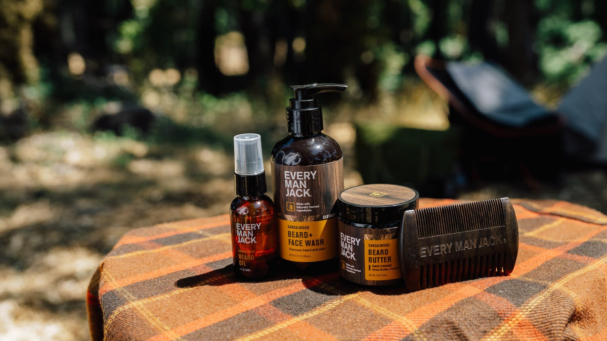 Every Man Jack beard-care products (beard oil, beard + face wash, beard butter) and wooden comb on plaid blanket