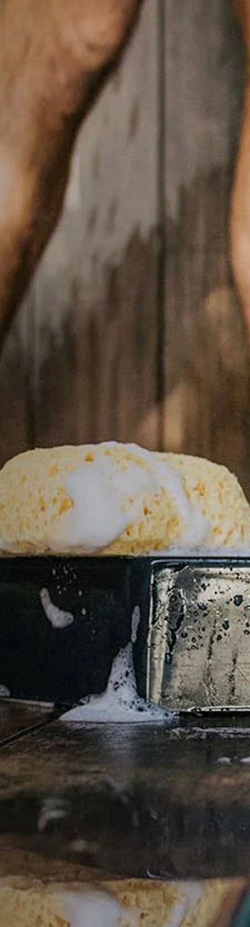 Three Every Man Jack body wash bottles labeled Sandalwood, Sea Salt, and Cedarwood with soap suds and a sponge on a wet wooden floor, with a person's legs in the background.