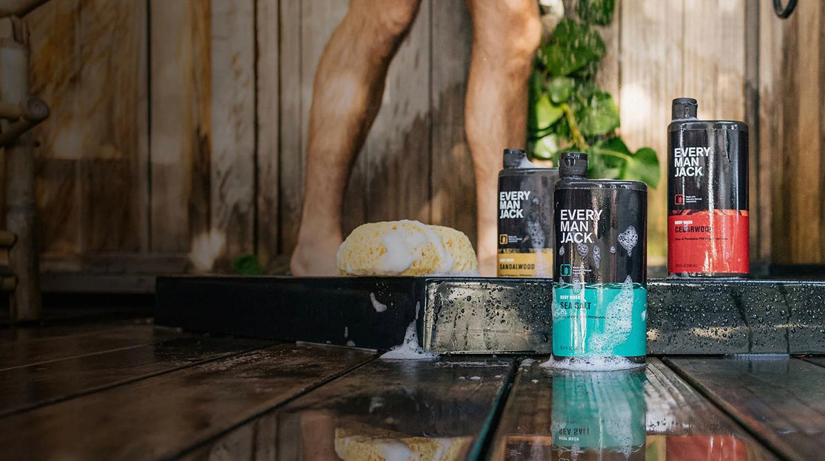 Three Every Man Jack body wash bottles labeled Sandalwood, Sea Salt, and Cedarwood with soap suds and a sponge on a wet wooden floor, with a person's legs in the background.