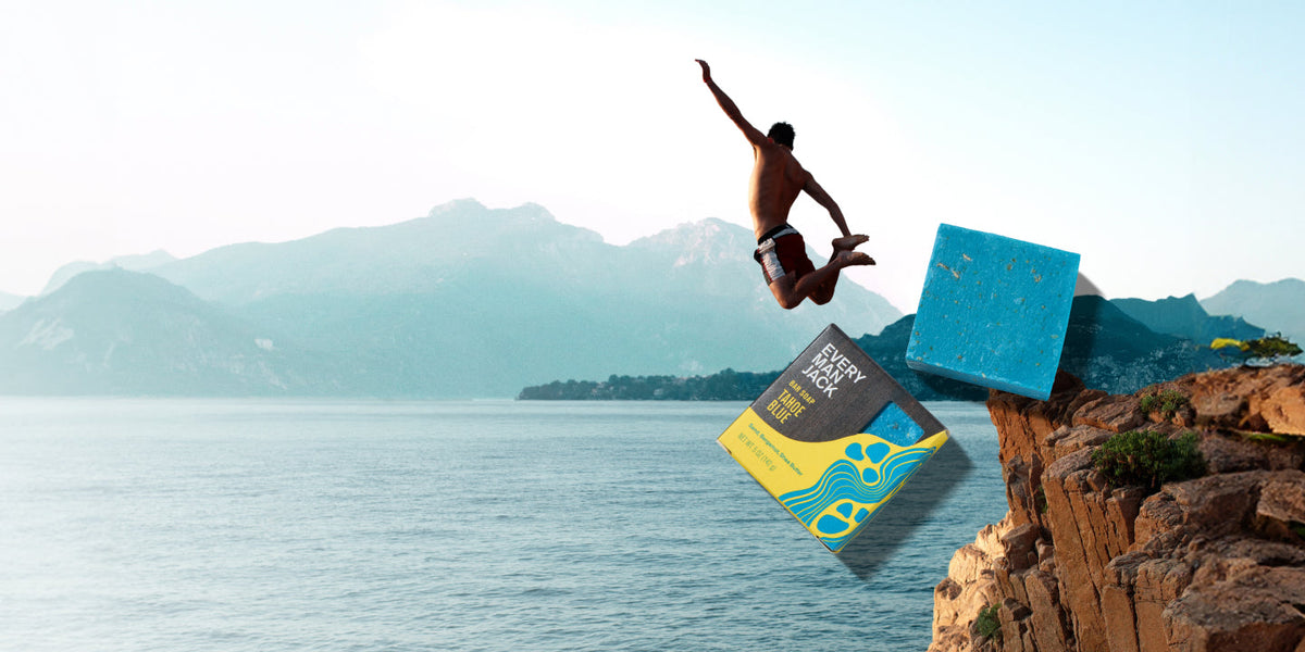 Man jumping off a rocky cliff into the ocean with a bright blue Every Man Jack bar soap and its yellow and blue box floating beside him against a mountain coastline