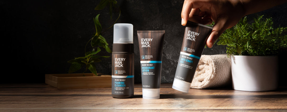Front view of three Every Man Jack Below the Belt products with hand picking one on wooden counter, towel and plant