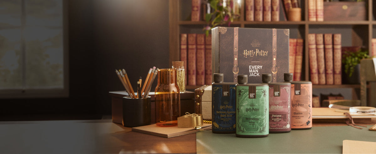 Four Harry Potter themed Every Man Jack body care bottles displayed on a desk in front of a Harry Potter trunk and bookcase with warm study lighting