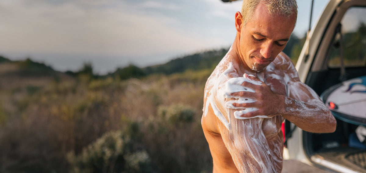 Man applying foamy body wash to his shoulder beside an open SUV with a surfboard on a coastal trail