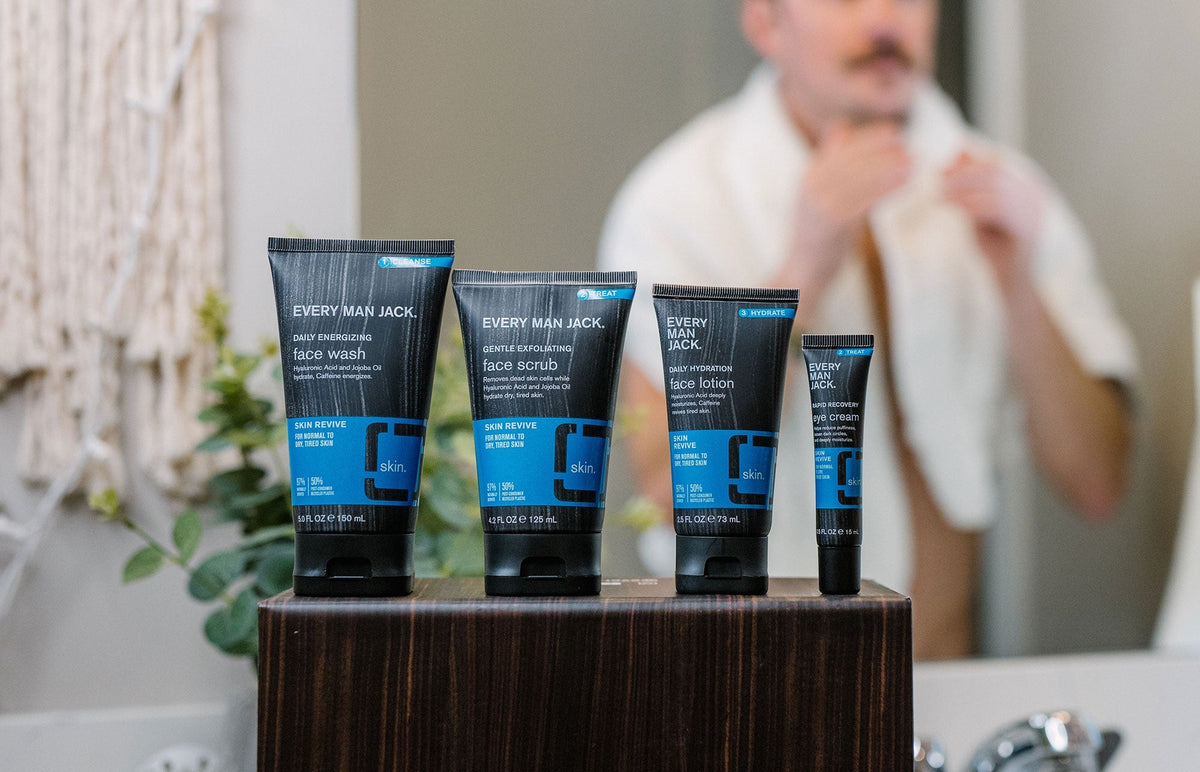 Four Every Man Jack face care tubes arranged on a wooden stand in a bathroom with a man adjusting a towel in the mirror behind them