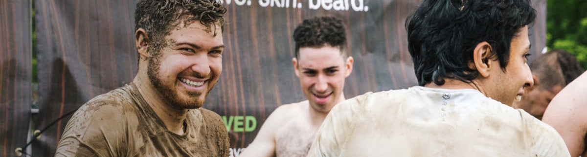 Three men laughing and covered in mud at an outdoor team event in front of a wood backdrop