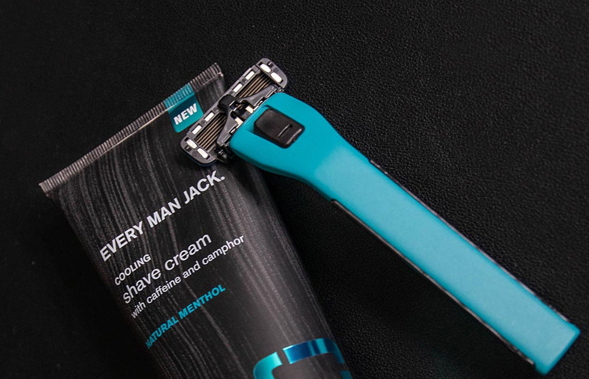 Angled view of a teal-handled razor and Every Man Jack cooling shave cream tube on a black textured background