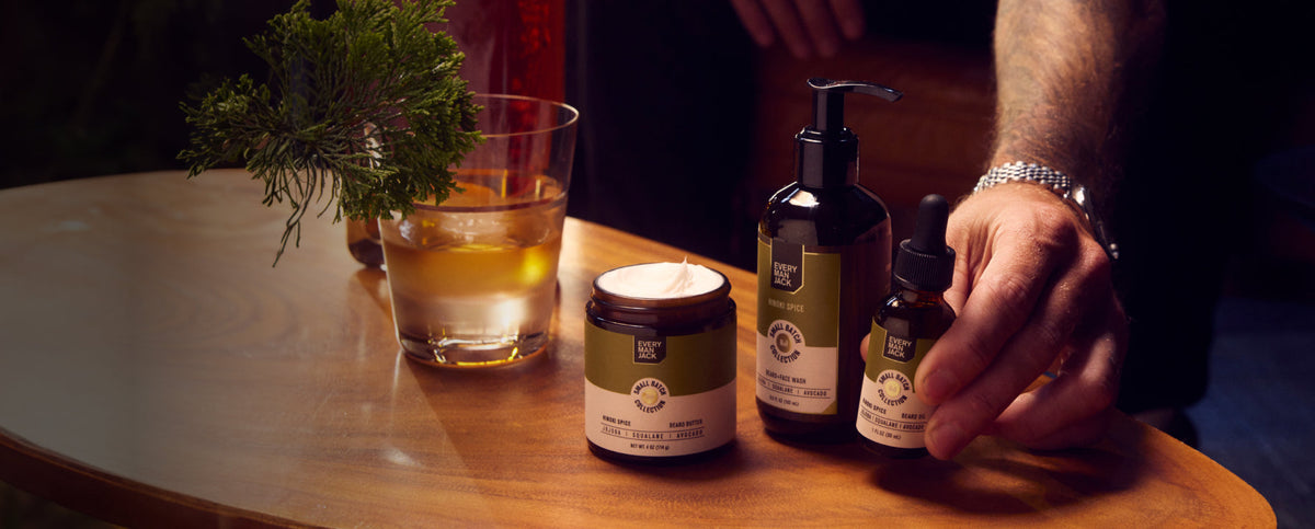 Hand reaching for a beard oil bottle among beard butter and face wash on a wooden table with a glass and greenery in warm light