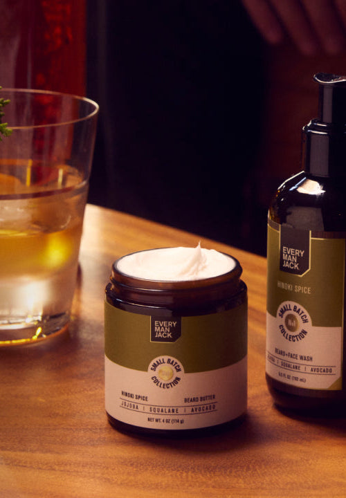 Hand holding a bottle of Every Man Jack Small Batch Collection Beard Oil, next to Beard Butter and Beard Face Wash on a wooden table with a glass of drink and greenery. 