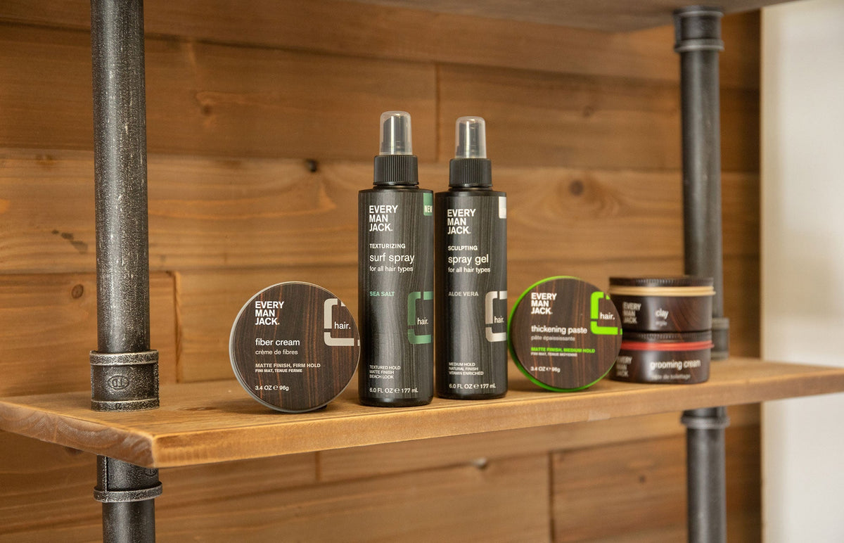 Every Man Jack hair sprays and styling jars arranged on a wooden shelf with a warm wood-paneled background