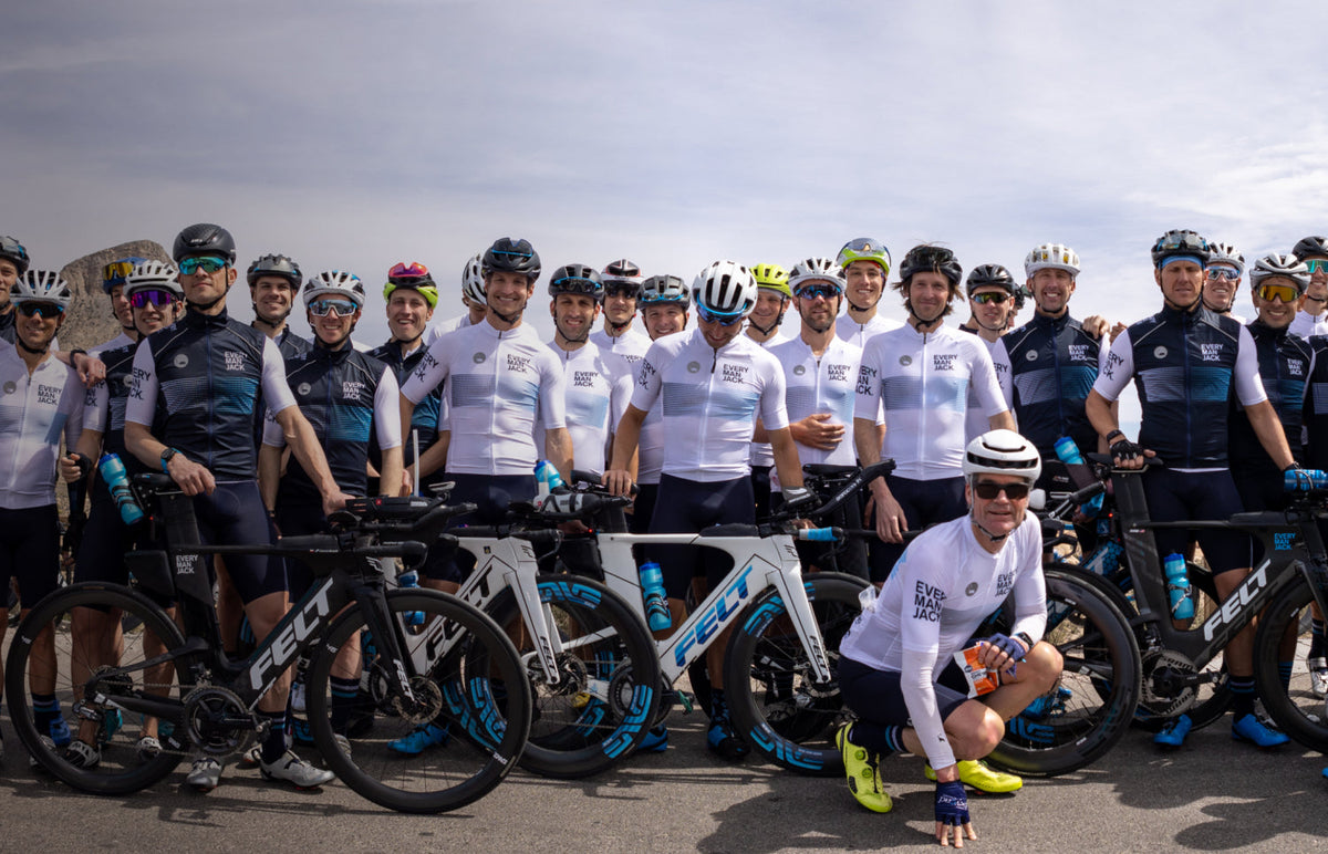 Every Man Jack triathlon team posing with road bikes on a road, wearing matching white and navy jerseys and helmets
