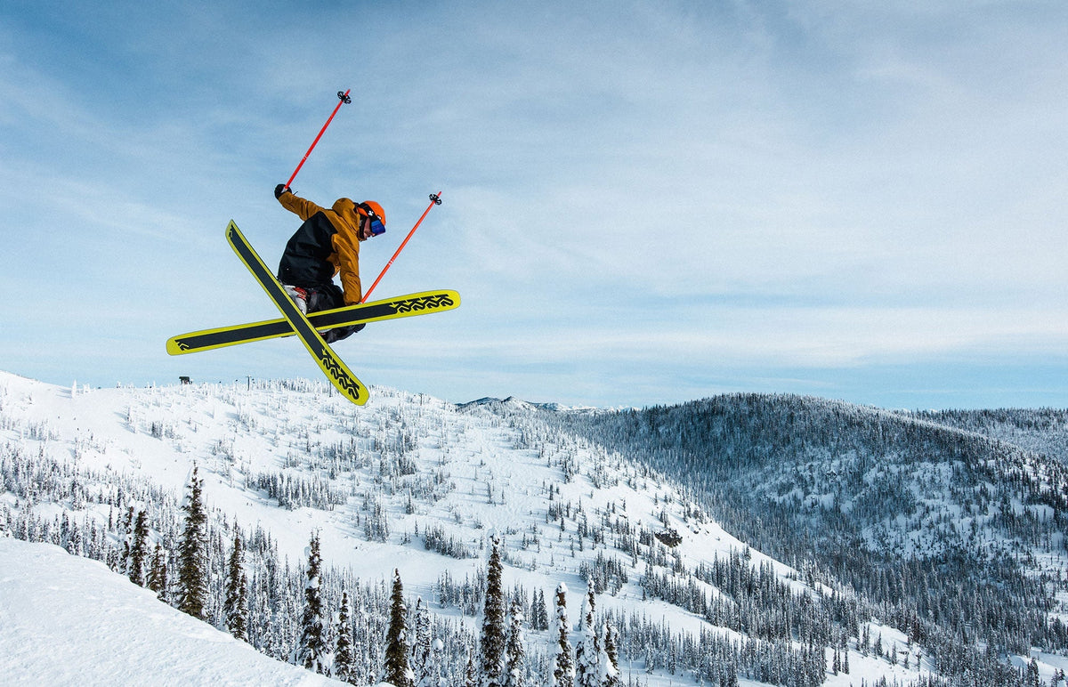 Skier launching into a jump over snow-covered mountain slopes with pine trees and a pale blue sky