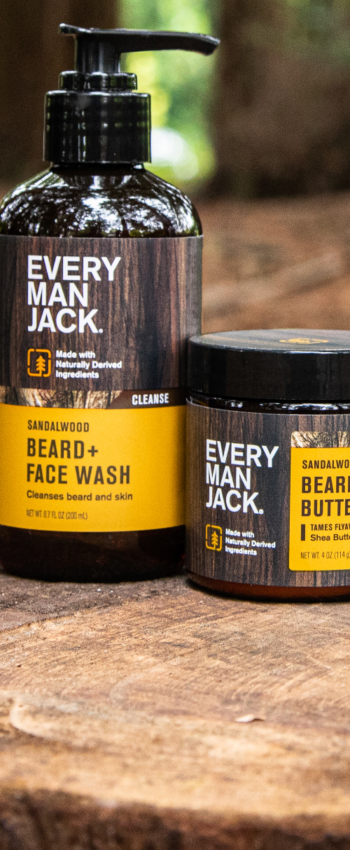 Every Man Jack beard care products on a tree stump: Sandalwood Beard + Face Wash, Beard Butter, Beard Oil, and a wooden comb with Every Man Jack engraved.