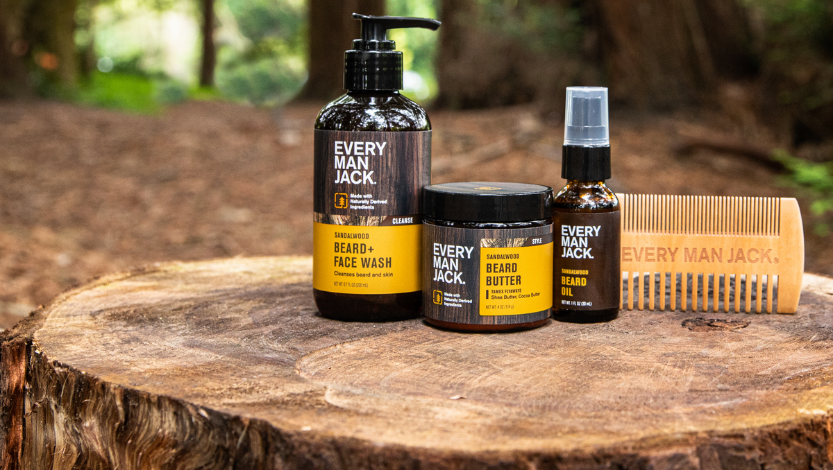 Every Man Jack beard care products on a tree stump: Sandalwood Beard + Face Wash, Beard Butter, Beard Oil, and a wooden comb with Every Man Jack engraved.