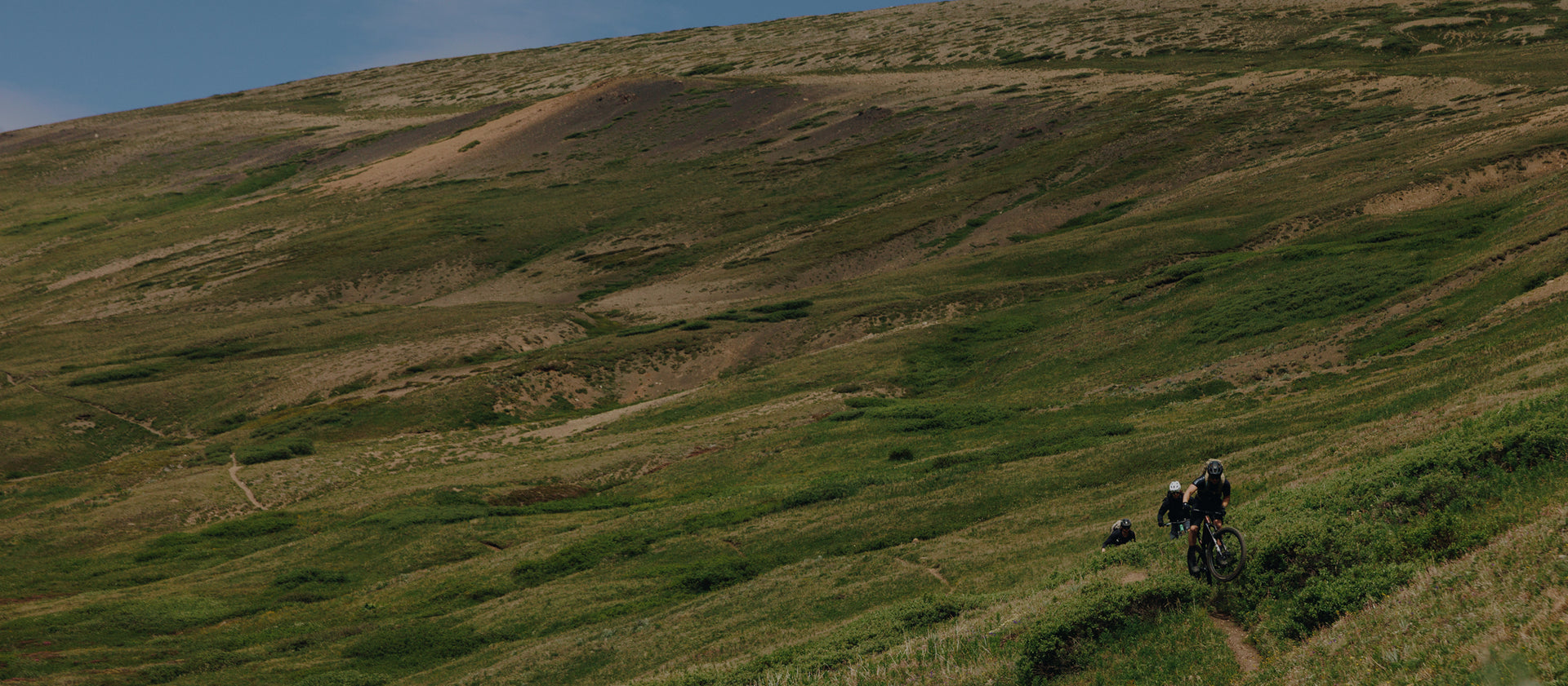 Three mountain bikers riding up a narrow dirt trail across a wide grassy hillside