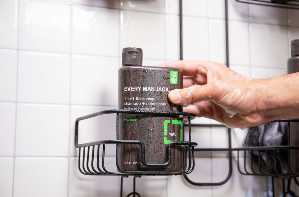 Hand reaching for an Every Man Jack 2-in-1 Thickening Shampoo and Conditioner, Tea Tree, in a wet shower caddy