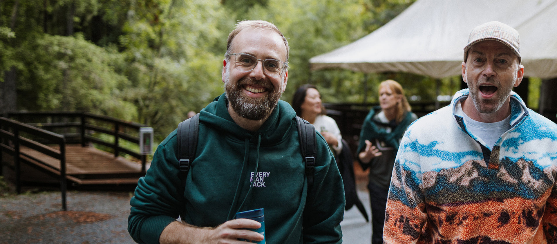 Smiling man in an Every Man Jack hoodie holds a travel mug on a forested trail