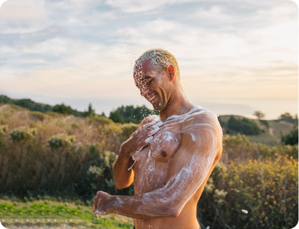 A man applying shampoo outdoors, with suds on his chest and a scenic background.