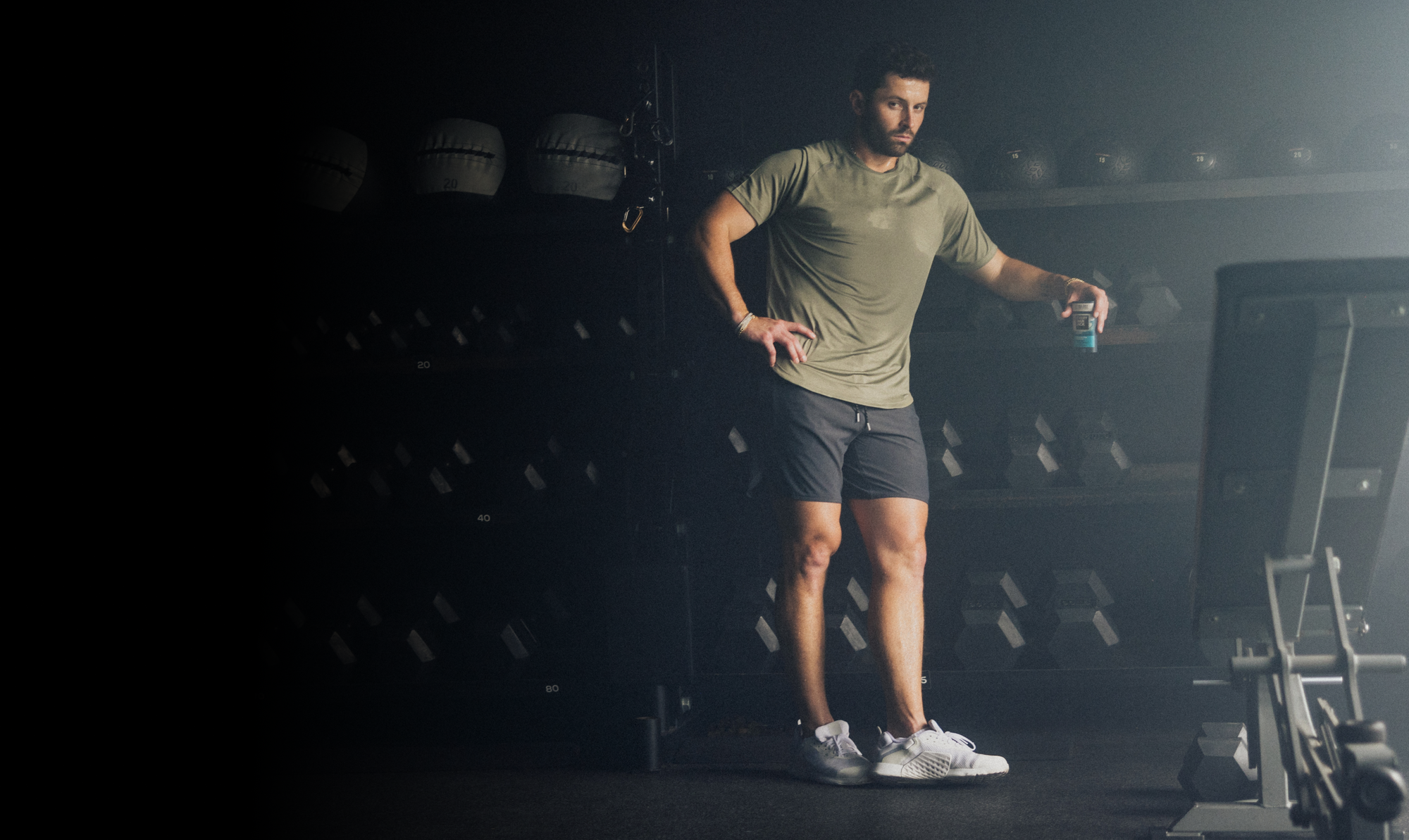 Baker Mayfield in a gym wearing an olive green athletic t-shirt and dark gray shorts, holding a spray bottle.