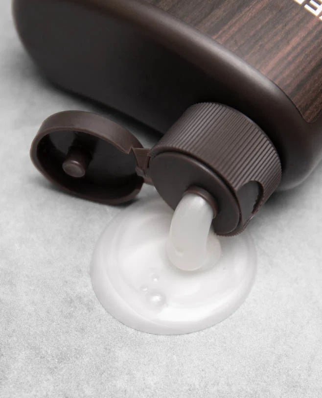 Close-up of 2-in-1 Thickening Shampoo + Conditioner bottle dispensing white shampoo from open flip-top cap on gray surface