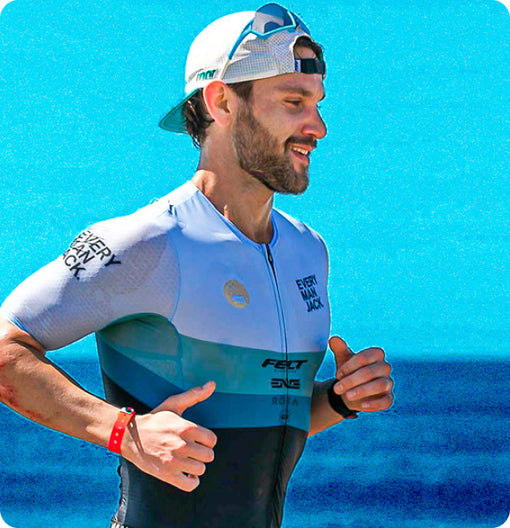 A man running in a triathlon suit with 'Every Man Jack' branding, smiling against a blue ocean background.