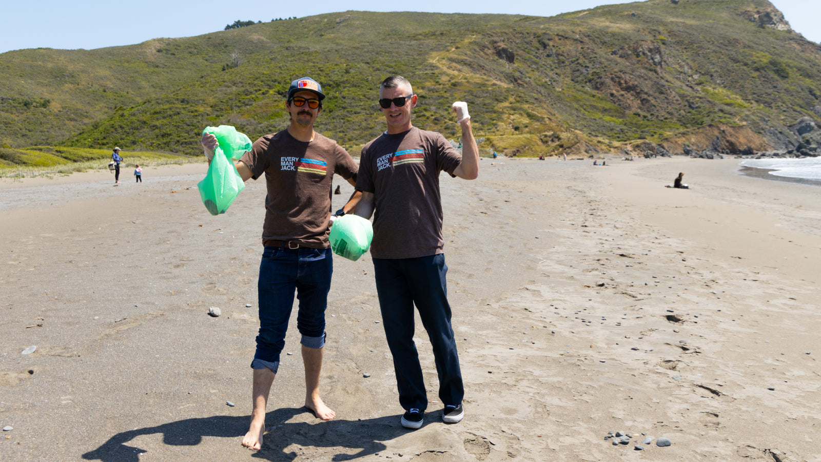 Two men in brown shirts holding green trash bags on a beach with hills in the background.