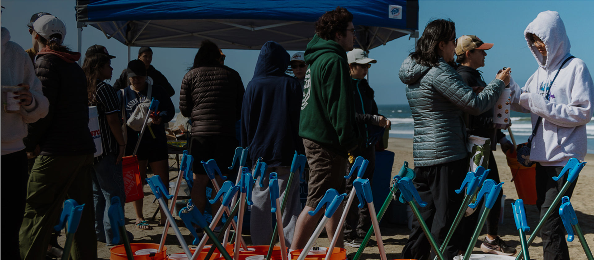 Volunteers cleaning a beach with orange buckets and blue-handled litter pickers under a canopy, ocean in background