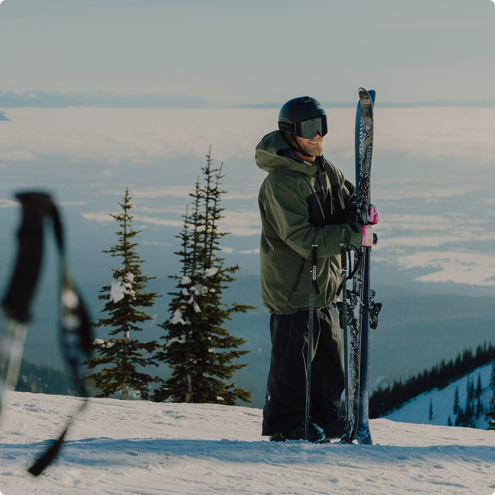 Man holding skis and poles standing on a snowy mountain ridge with pine trees and a cloud-filled valley below