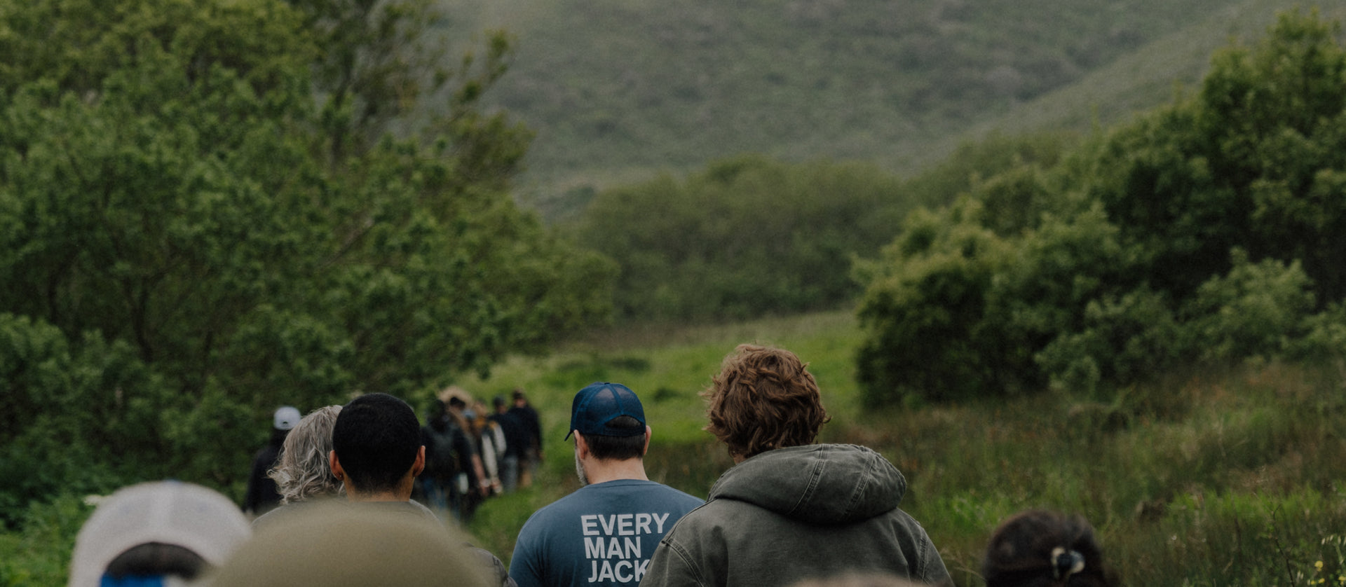 Group of people walking on a grassy trail toward green hills, one wearing a blue T‑shirt reading EVERY MAN JACK