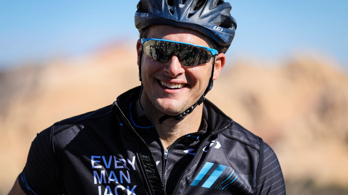 A smiling cyclist in a branded Every Man Jack jersey and sunglasses, outdoors.