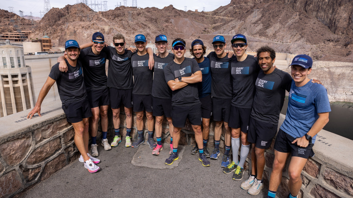 A group of 12 male athletes wearing matching Every Man Jack shirts and hats, posing together at a scenic location.